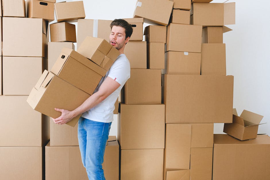 A man with dark hair and a light complexion is seen wearing a white t-shirt and blue jeans while inside a room, lifting a cardboard box with both arms. He appears to be engaged in a home relocation or furniture transport process, surrounded by numerous stacked cardboard boxes of varying sizes, some sealed with packing tape and others partially open. The boxes are arranged against a plain white wall, with some placed directly on the floor and others stacked on top of each other. The scene suggests an active packing or unpacking phase, possibly with protective materials like bubble wrap or blankets, as part of a detailed moving service. A moving trolley or hand truck might be nearby but is not visible in the image. The environment is well-lit, typical of indoor lighting, supporting the logistics of packing and moving activities. This image is relevant to [COMPANY_NAME]'s removals services focused on residential moves in Millbank, reflecting careful handling and organisation during the loading process for house relocation.