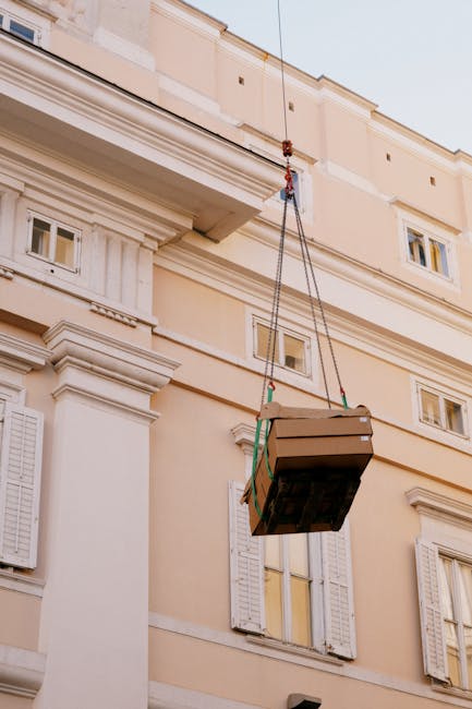 A large cardboard box secured with green straps is suspended in mid-air by a lifting apparatus connected to a crane or hoist, being lowered or hoisted outside a cream-colored residential building with white decorative cornices and window shutters. The box is positioned near the building's exterior wall, close to a window with yellow curtains. The scene takes place on a bright day, with natural light illuminating the facade, and the box appears to be part of a home relocation or furniture transport process, as part of a house removals service by Man with Van Millbank. The image captures the logistical aspect of loading or unloading household items, with the box clearly visible against the building's classic architectural details.