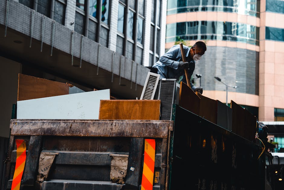 A worker wearing a face mask and casual clothing, including a light blue shirt and dark trousers, is standing inside the flatbed of a large, open-top black skip truck during daytime. The truck is parked on a city pavement in front of modern, multi-storey office buildings with glass and metal facades. Inside the skip, there are various household items and bulky waste materials, including wooden furniture panels, white and brown cupboard doors, plastic window frames, and wrapped packages, arranged in an upright and leaning position. The worker is lifting and positioning a piece of furniture or discarded household item using one hand, while holding a tool or strap with the other. The scene depicts the loading process involved in home removals or waste clearance, with natural daylight illuminating the urban environment, and the setting emphasizing the logistics of furniture transport and disposal for house removals, as managed by companies like Man with Van Millbank.