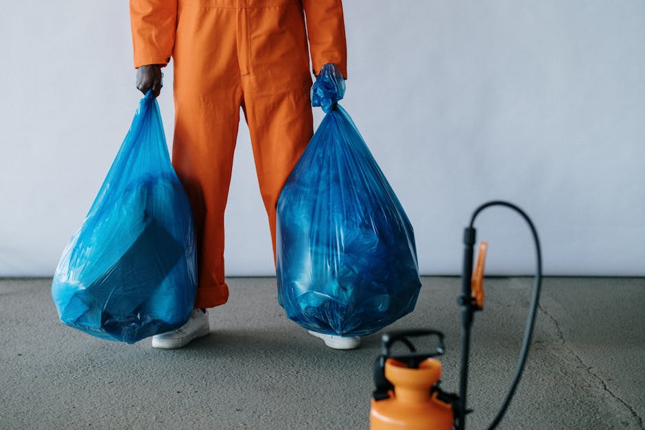 A person dressed in orange overalls is standing indoors on a plain grey floor, holding two large blue plastic bags filled with waste or recyclable materials. The person's feet are visible, wearing white footwear, while their hands grip the handles of the bags, indicating they are preparing for waste removal or disposal as part of house clearance or packing during a home relocation. In the foreground, a small section of an orange handheld sprayer or cleaner is visible, and the background consists of a plain, light-colored wall with soft, even lighting that highlights the movement and materials involved. This scene reflects the process of loading waste or bulky debris, possibly associated with a professional removals service like Man with Van Millbank, assisting with furniture transport, packing, or clearance in the context of a house move or removal project.