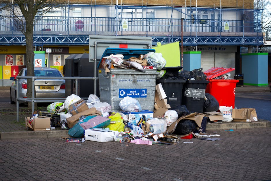 A worker wearing a face mask and casual clothing, including a light blue shirt and dark trousers, is standing inside the flatbed of a large, open-top black skip truck during daytime. The truck is parked on a city pavement in front of modern, multi-storey office buildings with glass and metal facades. Inside the skip, there are various household items and bulky waste materials, including wooden furniture panels, white and brown cupboard doors, plastic window frames, and wrapped packages, arranged in an upright and leaning position. The worker is lifting and positioning a piece of furniture or discarded household item using one hand, while holding a tool or strap with the other. The scene depicts the loading process involved in home removals or waste clearance, with natural daylight illuminating the urban environment, and the setting emphasizing the logistics of furniture transport and disposal for house removals, as managed by companies like Man with Van Millbank.