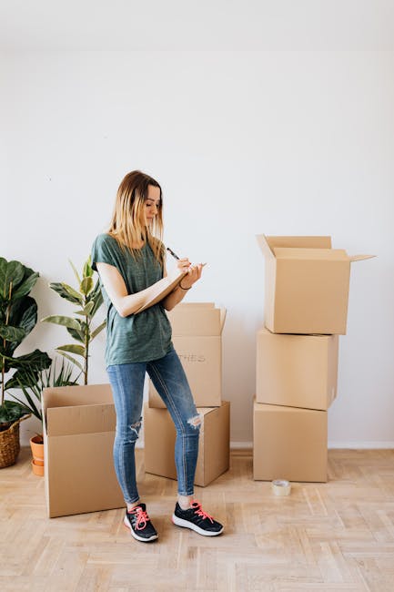 A young woman with shoulder-length blonde hair, dressed in a green t-shirt, ripped blue jeans, and black sneakers with pink laces, stands inside a room with light-colored wooden flooring and a plain white wall. She is holding a clipboard and pen, appearing to take notes as part of the packing process for a house move. Surrounding her are several cardboard boxes of various sizes, some closed and others open with flaps lifted, ready for packing or transport. To her left, there is a large potted plant with broad green leaves, placed on the floor next to a smaller plant in an orange pot. The scene suggests an early stage of home relocation, with ready-to-move packing materials and furniture, and the image is associated with professional removals and furniture transport services offered by Man with Van Millbank during a short-distance move from Horseferry Road to John Islip, aligned with the page title 'Horseferry Road to John Islip: Short-Move Checklist, MILLBANK'.