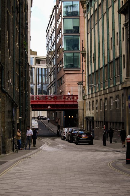 A narrow city street in central London, lined with historic stone buildings on the left and modern glass skyscrapers in the background. The street is closed to traffic with metal barriers and features a designated bicycle lane marked with white painted lines and a bicycle symbol. There are parked bicycles on the sidewalk and a few pedestrians visible, some walking along the pavement. The scene appears to be overcast with grey skies, and the overall environment reflects an urban setting suitable for house and office removals and furniture transport. Man with Van Millbank may coordinate moving services in such busy city streets, ensuring safe loading and unloading of household items attached to buildings and vehicles nearby.
