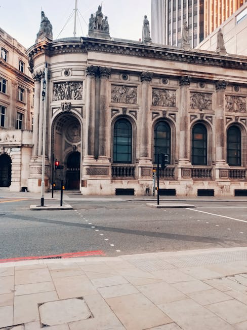 A historic, ornate stone building with large arched windows and detailed sculptures on the upper corners, situated on a city street with traffic lights and bollards in the foreground. The building has classic architectural features, including decorative stone carvings and columns. The surrounding environment includes modern high-rise structures in the background, and the street is lined with pavement and roadside markings. This setting suggests a central urban area suitable for house removals or furniture transport services. Man with Van Millbank, a professional removal company, may utilize such locations for loading or unloading during home relocation or furniture moving tasks, emphasizing efficient logistics and precise site handling.