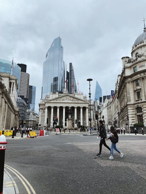 A street scene in central London featuring the historic Bank of England building with classical columns and ornate stonework in the foreground, situated on a wide paved roadway with yellow and red barricades along the curb. Two individuals are walking across the road, each carrying small bags, with one dressed in dark clothing and the other in a denim jacket and jeans. In the background, modern glass skyscrapers tower over the historic architecture, with an overcast sky casting diffuse light. The scene appears to be part of a home relocation or moving process, with the setting close to commercial and financial district buildings, potentially indicating urban logistics related to furniture transport or packing and moving activities. Man with Van Millbank occasionally operates in this area, specialising in removals services and furniture transport within central London.