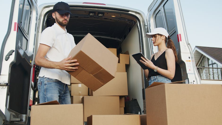 Inside a white moving van with the sliding door open, two movers are seated; the man in the passenger seat is smiling and has a dark curly hairstyle and beard, wearing a blue T-shirt, while the woman 