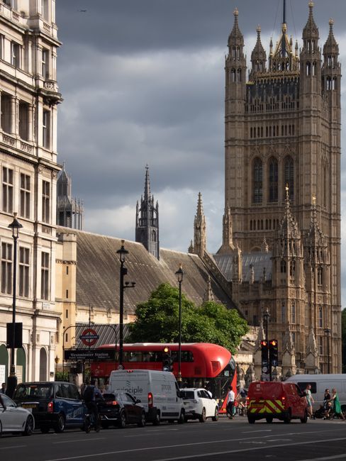 A street scene in Westminster, showing a busy road with various vehicles including a white van, a red double-decker bus, a red emergency vehicle, and several cars. A group of people is walking along the pavement, some carrying bags or boxes. In the background, the historic architecture of the Palace of Westminster with its Gothic-style towers and detailed stonework is visible, set against a cloudy sky. On the left side, part of a white building with decorative windows and a shopfront can be seen, with a black lamp post nearby. The scene captures the urban environment typical for house removals and furniture transport activities, with [COMPANY_NAME] providing professional moving services in such city locations.