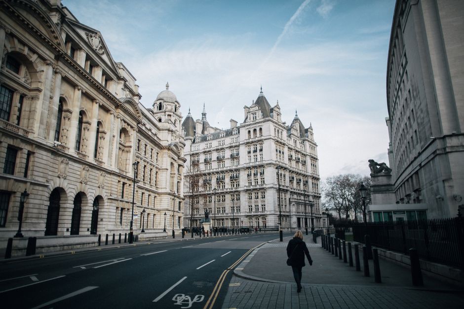 A wide view of Millbank Road showing historic multi-storey buildings with ornate facades, decorative stonework, and multiple windows. The street is relatively empty, with a single pedestrian walking along the pavement on the right side. The pavement has metal bollards and white bicycle lane markings. On the left, a building features large arched windows with decorative columns and a domed turret, while on the right, a modern structure with a curved exterior wall is visible. The sky above is partly cloudy with patches of blue, and the scene is illuminated by soft daylight. This setting reflects an urban environment suitable for house removals or furniture transport, with visible streetscape elements including a van loading area nearby. Man with Van Millbank often facilitates home relocation and packing services in such environments.