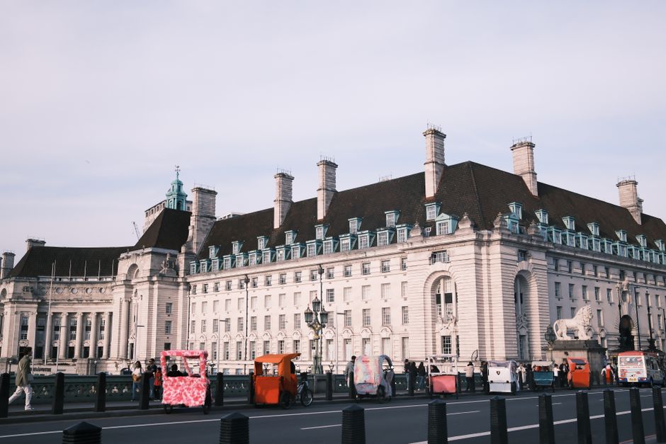 A street scene in Westminster, showing a busy road with various vehicles including a white van, a red double-decker bus, a red emergency vehicle, and several cars. A group of people is walking along the pavement, some carrying bags or boxes. In the background, the historic architecture of the Palace of Westminster with its Gothic-style towers and detailed stonework is visible, set against a cloudy sky. On the left side, part of a white building with decorative windows and a shopfront can be seen, with a black lamp post nearby. The scene captures the urban environment typical for house removals and furniture transport activities, with [COMPANY_NAME] providing professional moving services in such city locations.