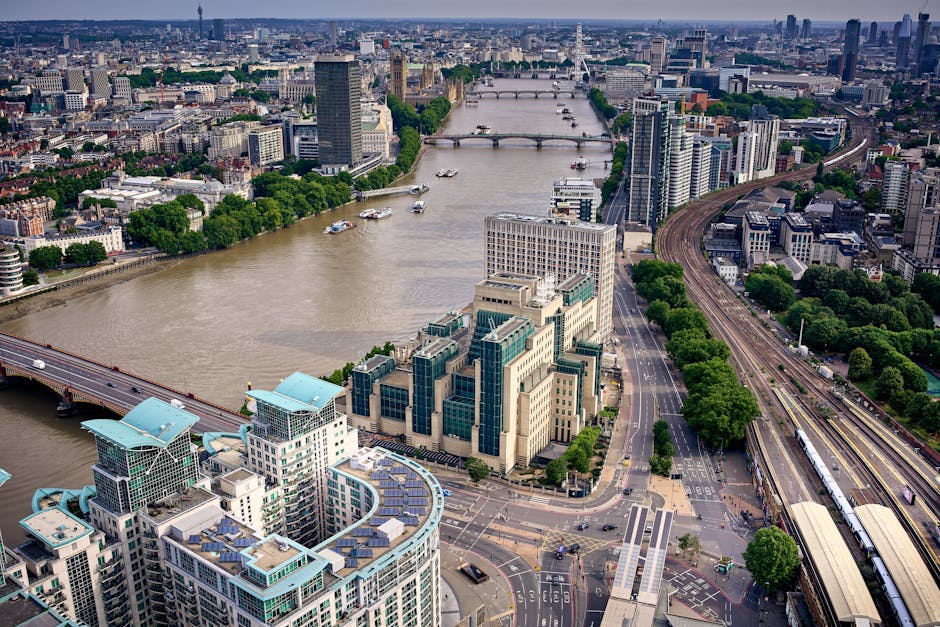 An aerial view of the River Thames in London, with several bridges crossing the waterway and a skyline featuring a mix of modern high-rise buildings and historic structures. Alongside the river, there are trees lining the streets and open spaces, with busy roads and railway lines visible. In the foreground, a group of buildings with distinctive glass and concrete facades are situated near the riverbank, indicative of urban development focused on commercial and residential use. The scene captures the transportation infrastructure, including bridges and railway tracks, supporting city logistics and home relocations. This setting is relevant for house removals, showcasing the dense urban environment where [COMPANY_NAME] provides moving and furniture transport services as part of efficient packing and loading processes during home relocation in the Millbank area. The overall lighting indicates daytime with natural sunlight illuminating the cityscape, highlighting the logistical complexity of residential moves within this busy, accessible city environment.