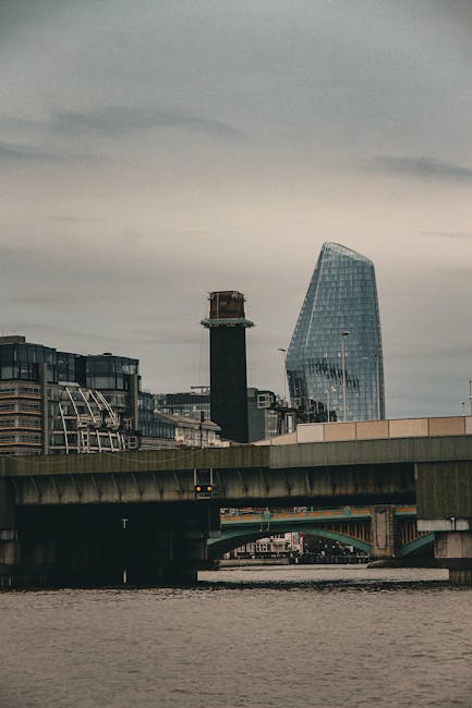 A view of London’s Millbank area showing a concrete bridge crossing over the River Thames in the foreground with calm water and a cloudy sky above. In the background, there are modern commercial and office buildings, including a distinctive curved glass skyscraper and a dark, rectangular tower under construction with scaffolding around its upper part. The scene captures an urban landscape typical of a house removal or furniture transport process in the area. The image’s natural lighting and perspective highlight the architectural features and the busy city environment, aligning with the services provided by Man with Van Millbank for home relocation and furniture moving tasks.