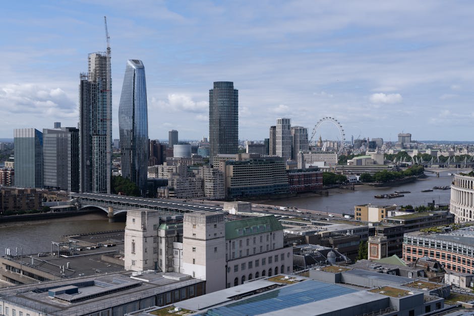 An aerial view of the River Thames in London, with several bridges crossing the waterway and a skyline featuring a mix of modern high-rise buildings and historic structures. Alongside the river, there are trees lining the streets and open spaces, with busy roads and railway lines visible. In the foreground, a group of buildings with distinctive glass and concrete facades are situated near the riverbank, indicative of urban development focused on commercial and residential use. The scene captures the transportation infrastructure, including bridges and railway tracks, supporting city logistics and home relocations. This setting is relevant for house removals, showcasing the dense urban environment where [COMPANY_NAME] provides moving and furniture transport services as part of efficient packing and loading processes during home relocation in the Millbank area. The overall lighting indicates daytime with natural sunlight illuminating the cityscape, highlighting the logistical complexity of residential moves within this busy, accessible city environment.
