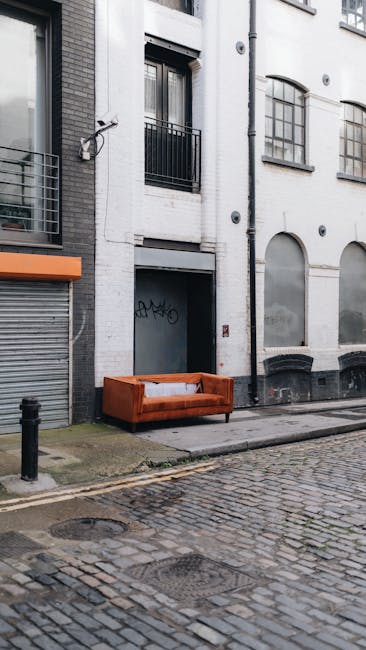 A photograph of a street scene outside a white residential building on Thorney Street, Millbank, showing a vintage orange sofa placed on the sidewalk in front of the building's entrance. The building has arched and rectangular windows, some with black metal railings and a small balcony, and a grey metal door beneath them. The cobblestone street features a curb and a black bollard, with the sidewalk made of concrete slabs. There are no visible people or vehicles, and the scene is illuminated by natural daylight. This outdoor setting may be part of a home relocation or furniture transport process coordinated by Man with Van Millbank, with the sofa possibly awaiting collection or delivery as part of a packing and moving operation.