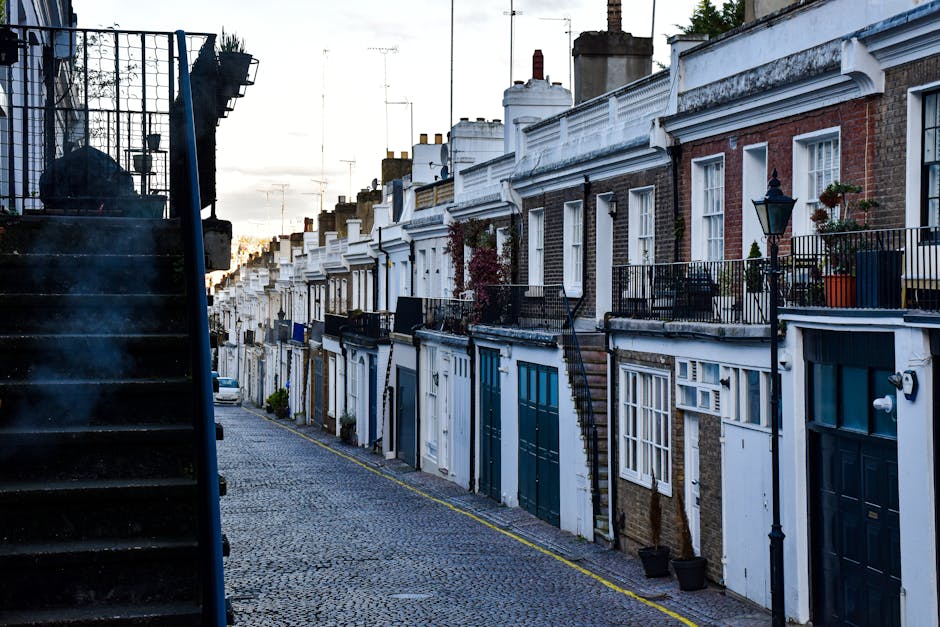 An exterior view of a narrow cobbled street in Thorney Street, Millbank, featuring a row of terraced houses with varying brick and white-painted facades. Each house has a small front balcony with black metal railings and potted plants, along with large double-glazed windows. On the left, a dark metal staircase with a black railing ascends alongside the buildings, partially visible at the edge of the image. The pavement is lined with a yellow border, and several small potted plants are placed near the entrances. The scene is captured during daylight hours with overcast sky, providing natural, diffused lighting. This setting exemplifies typical London residential architecture, often associated with home relocations where careful packing and furniture transport are essential, as undertaken by professional moving services such as Man with Van Millbank.