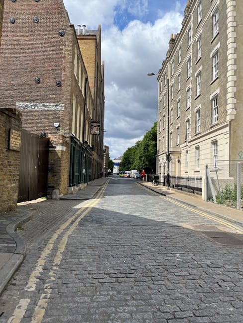 A photograph of a street scene outside a white residential building on Thorney Street, Millbank, showing a vintage orange sofa placed on the sidewalk in front of the building's entrance. The building has arched and rectangular windows, some with black metal railings and a small balcony, and a grey metal door beneath them. The cobblestone street features a curb and a black bollard, with the sidewalk made of concrete slabs. There are no visible people or vehicles, and the scene is illuminated by natural daylight. This outdoor setting may be part of a home relocation or furniture transport process coordinated by Man with Van Millbank, with the sofa possibly awaiting collection or delivery as part of a packing and moving operation.