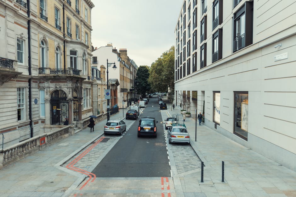 A view of a narrow urban residential street with a mix of classic and modern multi-storey buildings on either side. On the right, a modern cream-coloured building features large windows and a smooth f