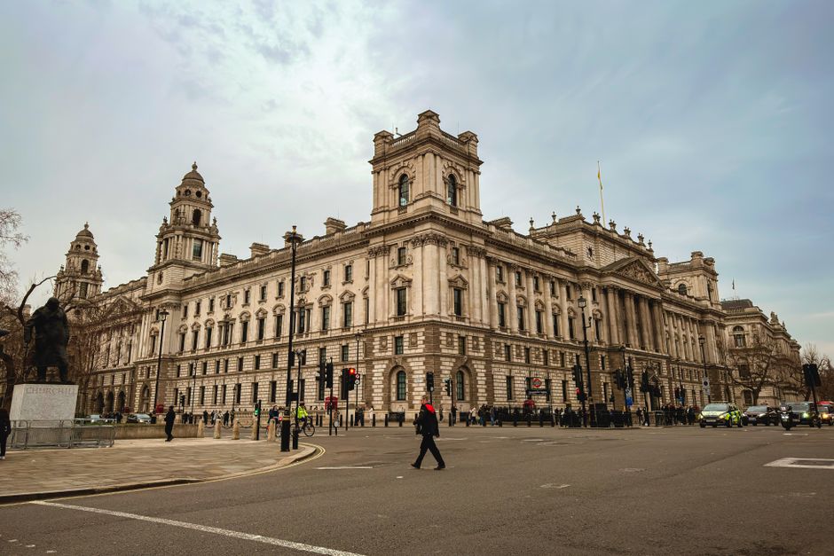 A large, historic stone building with ornate architectural details, including multiple towers and decorative window frames, situated at the corner of a busy street in central London. The building is illuminated by natural daylight under a cloudy sky, with pedestrians crossing the road and vehicles stopped at traffic lights nearby. Several people, some wearing high-visibility vests, are positioned on the pavement, possibly involved in moving or logistics activities. In the foreground, a man with a backpack walks across the street, while others stand near the building entrance. The scene captures an urban environment typical of a home relocation or furniture transport operation, with a focus on the surrounding architecture and the street activity in front of the historic structure, aligning with discussions on house removals and moving services like those provided by Man with Van Millbank.